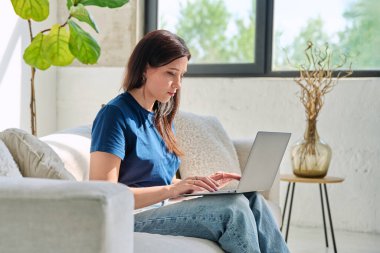 Genç, ciddi bir kadın dizüstü bilgisayarda oturuyor. Evdeki koltukta oturuyor. Bilgisayarı iş, eğlence, iletişim, alışveriş, serbest yazarlık, blog, eğitim için kullanan bir kadın. Teknoloji yaşam tarzı insan kavramı