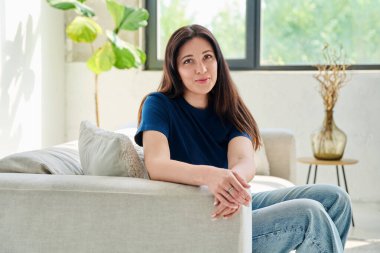 Oturma odasındaki kanepede oturan güzel, mutlu esmer kadının portresi. 30 'lu yaşlarda çekici bir kadın kameraya bakıyor. Güzellik, yaşam tarzı, kadın konsepti