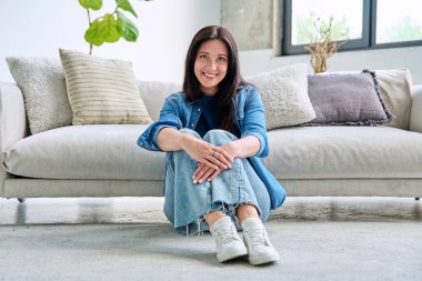 Oturma odasındaki kanepenin yanında oturan güzel, mutlu, esmer kadının portresi. 30 'lu yaşlarda çekici bir kadın kameraya bakıyor. Güzellik, yaşam tarzı, kadın konsepti