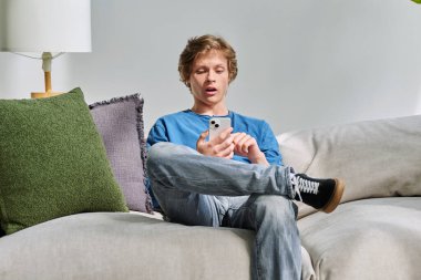 Evdeki kanepede oturan akıllı telefon kullanan yakışıklı, şaşırmış bir genç. Gençlik yaşı, ergenlik, yaşam tarzı, teknoloji konsepti