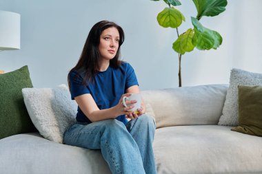 Oturma odasındaki kanepede oturan güzel, gülümseyen esmer kadının portresi. Mutlu, rahatlatıcı, çekici 30 'lu yaşlarda bir kadın başka tarafa bakıyor. Güzellik, yaşam tarzı, kadın konsepti