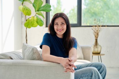 Oturma odasındaki kanepede oturan güzel, mutlu esmer kadının portresi. 30 'lu yaşlarda çekici bir kadın kameraya bakıyor. Güzellik, yaşam tarzı, kadın konsepti