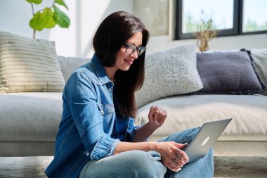 Genç bir kadın dizüstü bilgisayar ekranına bakıyor görüntülü sohbet görüşmesi yapıyor evde oturuyor, online toplantı yapıyor, uzaktan iletişim kuruyor, blog bloğu kaydediyor. Teknoloji işi insanları çalıştırır.