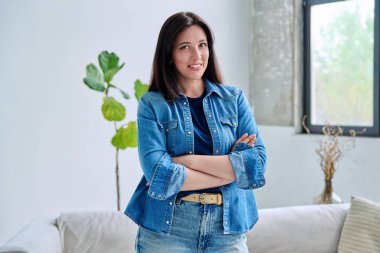 Evin içinde kendine güvenen güzel genç bir kadının portresi. Kollarını kavuşturup kameraya bakan kot elbiseli, gülümseyen kadın. Güzellik, yaşam tarzı, sağlık, 30 'ların insan kavramı