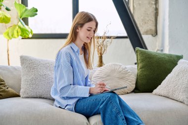 Evdeki kanepede dijital tablet kullanan genç bir kadın. Cihazlı kadın üniversite öğrencisi, rahatlama eğitimi, eğlence iletişimi eğitimi teknolojisi, yaşam tarzı gençlik konsepti