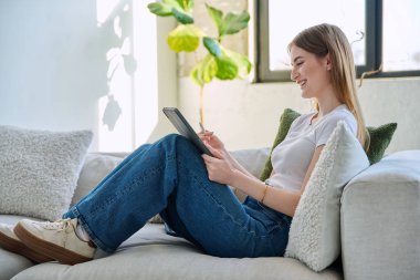 Genç ve güzel bir kız öğrenci dijital tabletle online sohbet konferansı yapıyor. Evde, kanepede oturuyor. İletişim öğrenme teknolojileri gençlerin video bloglarında boş zamanlarında çalışıyor