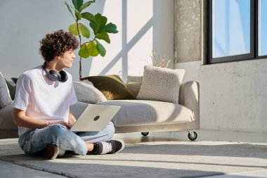 Evdeki kanepenin yanında dizüstü bilgisayarda yazı yazan yakışıklı bir erkek portresi, profil görüntüsü fotokopi alanı. Kıvırcık, güler yüzlü, 20 yaşında bir öğrenci. Yaşam tarzı eğitim teknolojisi gençlik kavramı