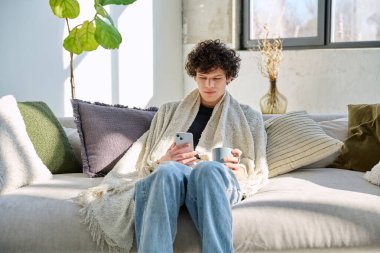 Sıcak bir battaniyenin altında, elinde bir kupa sıcak içecek ve akıllı telefonla evdeki oturma odasındaki kanepede oturan rahat genç bir adam. Mevsim sonbahar kışı, ev konforu yaşam tarzı rahatlığı