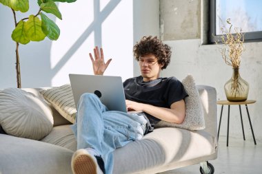 Genç yakışıklı kıvırcık adam internette sohbet etmek için bilgisayarını kullanıyor. Evde, koltukta oturuyor. İletişim öğrenimi için dijital teknolojiler boş zamanlarında sosyal medya bloglarında çalışır