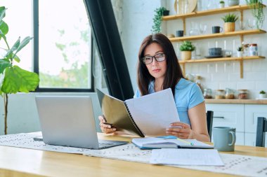 Dizüstü bilgisayarla çalışan, iş gazetelerini okuyan, mutfaktaki masada oturan ciddi bir kadın. Teknoloji, iş, iş, 30 'ların insan kavramı