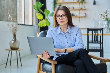 Orta yaşlı iş kadını dizüstü bilgisayarlı sandalyede oturuyor, 40 'lı yaşlarda kadın gülümsüyor oturma odasının iç tarafındaki kameraya bakıyor, psikolog terapist danışman koltukta uzman.