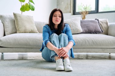 Evdeki kanepenin yanında oturan mutsuz genç kadın üzülüyor. Depresyon, stres, acı, sorunlar, akıl sağlığı insanları kavramı