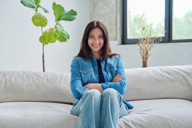 Oturma odasındaki kanepede oturan güzel, kendine güvenen esmer kadının portresi. 30 'lu yaşlarda çekici bir kadın çapraz kollarla kameraya bakıyor. Güzellik, yaşam tarzı, kadın konsepti