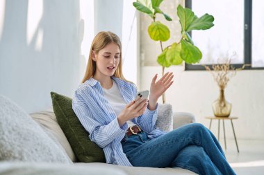 Genç ve güzel bir kadın video konferansı için akıllı telefonu kullanıyor. Evde, kanepede oturuyor. İletişim öğrenmek için dijital teknolojiler boş zamanlarında sosyal medya bloglarında çalışır
