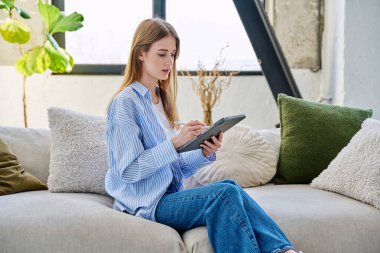 Evdeki kanepede dijital tablet kullanan genç bir kadın. Cihazlı kadın üniversite öğrencisi, rahatlama eğitimi, eğlence iletişimi eğitimi teknolojisi, yaşam tarzı gençlik konsepti