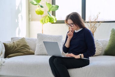 Evdeki kanepede oturup dizüstü bilgisayarda yazı yazan orta yaşlı güzel bir iş kadınının portresi, gevşemiş ciddi bir kadın. Teknoloji, yaşam tarzı, iş, eğlence, insan kavramı