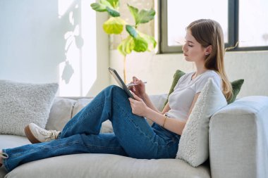 Evdeki kanepede dijital tablet kullanan genç bir kadın. Cihazlı kadın üniversite öğrencisi, rahatlama eğitimi, eğlence iletişimi eğitimi teknolojisi, yaşam tarzı gençlik konsepti