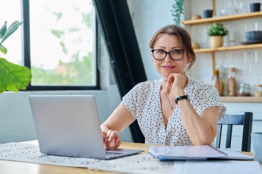 Orta yaşlı gülümseyen güzel bir kadın dizüstü bilgisayar belgeleriyle çalışıyor, mutfaktaki masada oturan kameraya bakıyor. Teknoloji, iş, iş, olgun insanlar konsepti