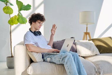 Genç yakışıklı kıvırcık adam internette sohbet etmek için bilgisayarını kullanıyor. Evde, koltukta oturuyor. İletişim öğrenimi için dijital teknolojiler boş zamanlarında sosyal medya bloglarında çalışır