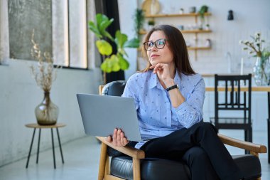 Orta yaşlı iş kadını dizüstü bilgisayarlı sandalyede oturuyor. Ev ofisinden 40 'lı yaşlarda kadın gülümsüyor. Oturma odasında.