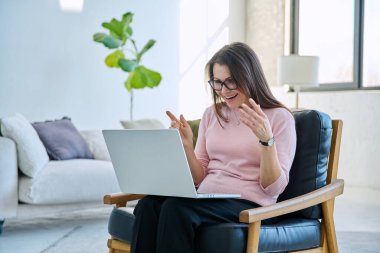 Orta yaşlı bir kadın uzaktan çalışıyor, video konferans görüşmeleri yapıyor, ofis koltuğunda dizüstü bilgisayar kullanıyor. Psikoloji oturumu çevrimiçi öğretim koçluğu vlog işi iletişimi