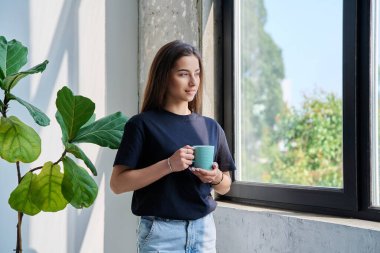 Güzel bir sükunetin portresi. Evde elinde fincanla dinlenen ve pencereden dışarı bakan rahat bir genç kız. Gençlik, yaşam tarzı, 17, 18, 19 yaş konsepti.