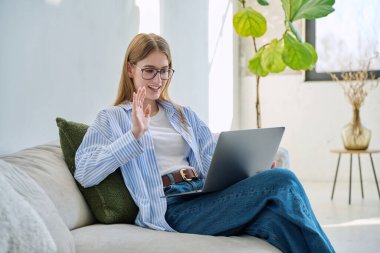 Genç kız video sohbet konferansı için dizüstü bilgisayar kullanıyor. Kanepede oturmuş web kamerasına bakıyor, bilgisayar ekranından konuşuyor. Eğitim eğitimi teknolojileri gençlik iletişimi kavramı üzerinde çalışır