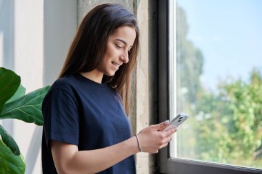 Akıllı telefonu kullanan genç bir kız pencerenin yanında duruyor, kopyalama alanı. Genç gülümseyen kız üniversite öğrencisi ekran kaydırma okumasına bakıyor, boş zamanlarında iletişim öğrenmek için mobil uygulamalar