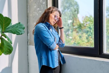 Olgun, üzgün, bunalımlı, stresli, zihinsel ve fiziksel acı çeken bir kadın pencerenin yanında duruyor. Sorunlar yalnızlık, baş ağrısı hormon stresi, orta yaşlı kadınların sağlığı