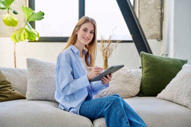 Dijital tablet kullanan genç bir kadın evdeki koltukta oturan kameraya bakıyor. Cihaz eğitimi alan bayan üniversite öğrencisi rahatlama eğitimi, eğlence iletişimi eğitimi teknolojisi, yaşam tarzı gençliği