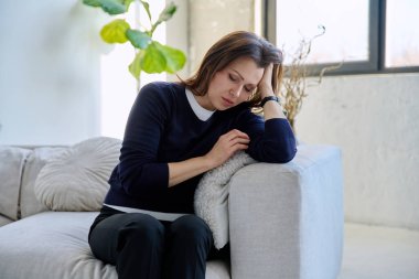 Olgun, üzgün, stresli bir kadın. Stresli, zihinsel ve fiziksel bir acı yaşıyor. Kanepede oturuyor. Sorunlar yalnızlık, baş ağrısı hormon stresi, orta yaşlı kadınların sağlığı