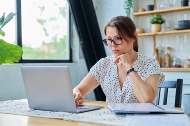 Orta yaşlı ciddi bir kadın dizüstü bilgisayar belgeleriyle çalışıyor, bilgisayarda daktilo yazıyor, mutfaktaki masada oturuyor. Teknoloji, iş, iş, olgun insanlar konsepti