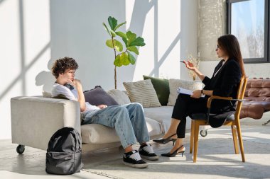 Genç erkek hasta, ofisinde oturan orta yaşlı kadın psikolog sosyal hizmet danışmanı ile konuşuyor. Gençlik psikoterapisi psikolojisi için ruh sağlığı psikolojik sosyal hizmetleri