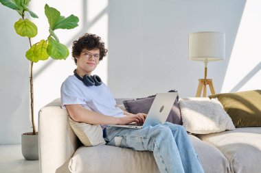 Portre yakışıklı, gülümseyen erkek dizüstü bilgisayar kullanıyor, kameraya bakıyor, evdeki kanepede oturuyor. 20 yaşında bilgisayar başında yazı yazan kıvırcık bir öğrenci. Yaşam tarzı eğitim teknolojisi gençlik kavramı