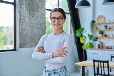 Evde çapraz kollarla poz veren kendinden emin başarılı genç kızın portresi. Kameraya bakan uzun saçlı, gülümseyen genç bir bayan. Güzellik, sağlık, yaşam tarzı, gençlik kavramı