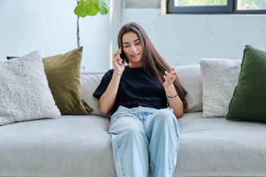 Genç, gülümseyen bir kadın evinde, koltukta oturuyor ve cep telefonuyla konuşuyor. Rahatlamış, güzel bir kadın. İletişim, ev konforu, yaşam tarzı, gençlik kavramı