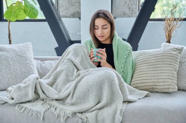Güzel genç bir kadın sıcak battaniyenin altında elinde fincanla oturma odasında, kanepede oturuyor. Soğuk bahar sonbahar kış mevsimi, tatiller, rahat, boş zaman, yaşam tarzı konsepti
