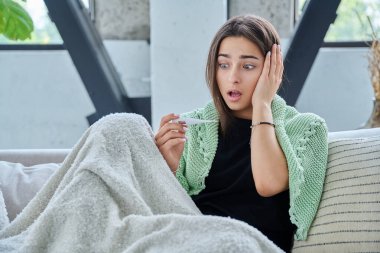 Üzgün genç kız evde battaniyenin altında oturan termometreye bakarken şaşırdı. Yüksek sıcaklıkta mevsimsel viral hastalıklar, hastalık, sağlık gençliği, ergenlik kavramı