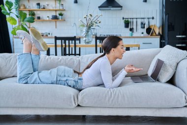 Genç kız video sohbet konferansı için dizüstü bilgisayar kullanıyor. Kanepede uzanmış web kamerasına bakıyor, bilgisayar ekranında konuşuyor. Eğitim eğitimi teknolojileri eğlence iletişimi gençlik kavramı