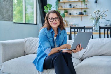Evdeki kanepede dizüstü bilgisayarı olan, rahat gülümseyen ve kameraya bakan orta yaşlı güzel bir kadının portresi. Teknoloji, yaşam tarzı, iş, eğlence, insan kavramı
