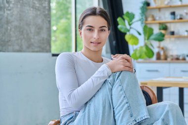 Evde poz veren güzel genç bir kadının portresi. 17-18 yaşlarında şık bir genç kız. Sandalyede oturmuş kameraya bakıyor. Gençler için güzellik, gençlik, yaşam tarzı, ürünler ve hizmetler