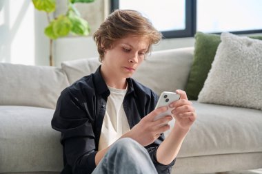 Evde oturmuş akıllı telefon kullanan ciddi yakışıklı bir genç. Teknolojiler, mobil başvurular boş zaman iletişim eğitimi uygulamaları