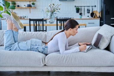 Evde dizüstü bilgisayarla kanepede uzanmış sınav için notlar alan genç üniversiteli kız. Eğitim e-öğrenim kursu online ders yaşam tarzı gençlik kavramı