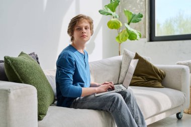 Yakışıklı, ciddi, genç bir çocuk evinde, kanepede oturuyor dizüstü bilgisayarında daktilo yazıyor ve kameraya bakıyor. Gençlik yaşı, ergenlik, yaşam tarzı, teknoloji konsepti