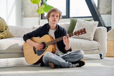 Akustik gitar laptopuyla yerde oturmuş kameraya bakan yakışıklı bir gencin portresi. Yaşam tarzı ergenlik eğitimi teknolojisi kavramı