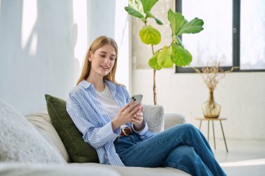 Kanepede oturan genç, gülümseyen bir kadın akıllı telefon kullanıyor. Rahat, güzel, doğal, elinde cep telefonu olan bir sarışın kadın. Boş zaman sosyal iletişim eğitimi teknolojileri çalışıyor