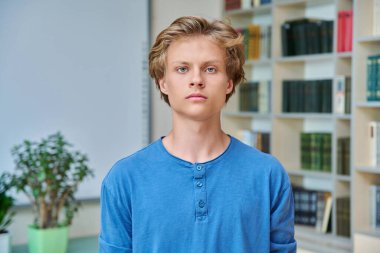 Kütüphanede kameraya bakan yakışıklı, ciddi bir gencin portresi. 16-17 yaşlarında genç bir erkek. Sınıfta kitap raflarıyla poz veriyor. Eğitim ergenlik bilgisi