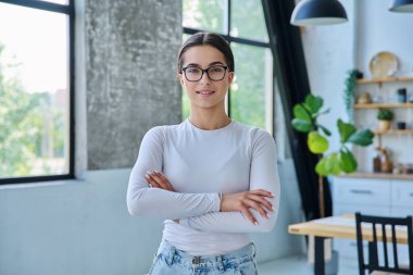 Evde çapraz kollarla poz veren kendinden emin başarılı genç kızın portresi. Kameraya bakan uzun saçlı, gülümseyen genç bir bayan. Güzellik, sağlık, yaşam tarzı, gençlik kavramı