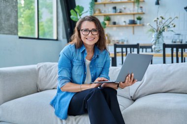Evdeki kanepede dizüstü bilgisayarı olan, rahat gülümseyen ve kameraya bakan orta yaşlı güzel bir kadının portresi. Teknoloji, yaşam tarzı, iş, eğlence, insan kavramı