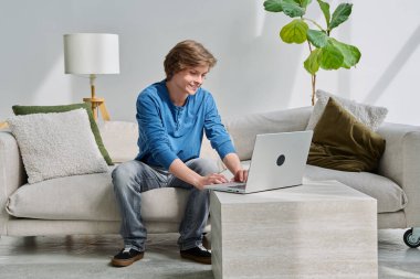 Yakışıklı, gülümseyen genç çocuk evinde, kanepede oturmuş dizüstü bilgisayarında daktilo yazıyor. Gençlik yaşı, ergenlik, yaşam tarzı, teknoloji konsepti